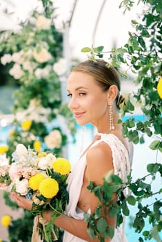 Bride with a bouquet of flowers stands half-turned near the wedding arch. High quality photo