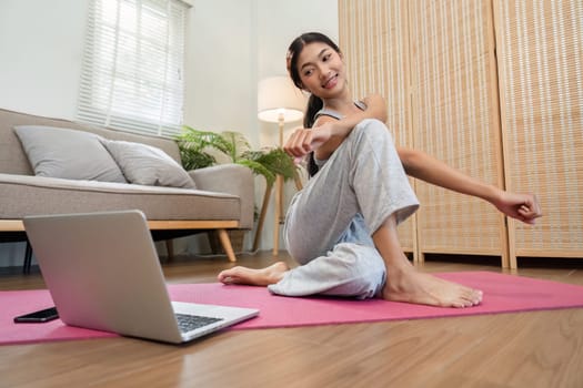 In a comfortable home setting, a woman engages in yoga stretching while using a laptop for online guidance.