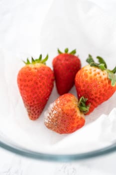 Bright red strawberries, interspersed with signs of mold, rest in a glass bowl lined with a paper towel on a white napkin, indicating improper storage techniques.