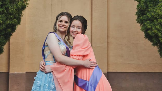 Two happy graduates in elegant dresses are hugging each other, celebrating their academic achievement and friendship
