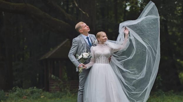 Newlyweds holding a bouquet and raising a long veil in a windy forest, celebrating their wedding day with joy and love