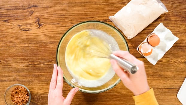 Flat lay. In a glass mixing bowl, a large hand whisk expertly blends the ingredients for a delectable gingerbread bundt cake with caramel frosting.