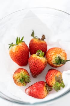 Bright red strawberries, interspersed with signs of mold, rest in a glass bowl lined with a paper towel on a white napkin, indicating improper storage techniques.