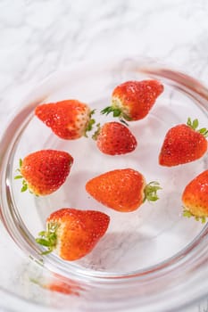 Ripe strawberries are submerged in water within a large glass mixing bowl, a step in washing the fruit to ensure cleanliness and longevity before storage or consumption.