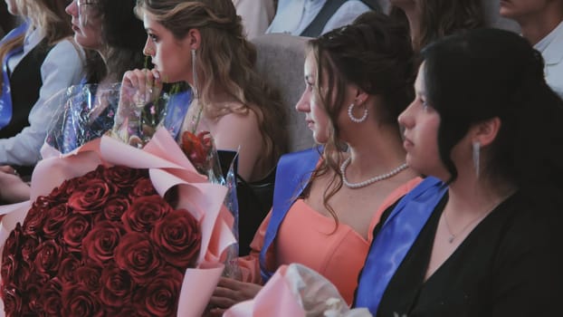 Female graduates attentively listening during graduation ceremony, clutching bouquets of vibrant red roses