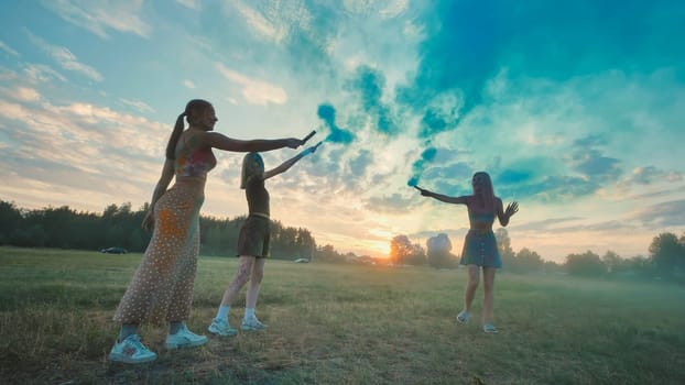 Three girls celebrating their friendship by playing with colored smoke bombs in a field at sunset