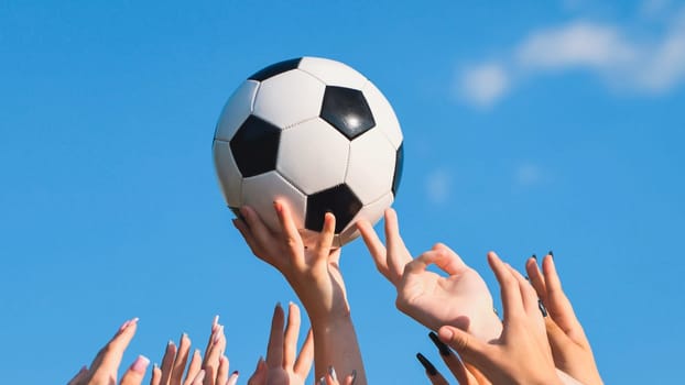 Numerous hands reaching for a soccer ball under a clear blue sky, representing teamwork, sportsmanship, and school pride