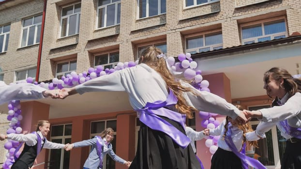 Group of happy students dancing together in a circle, celebrating their last school day with purple and white balloons and ribbons