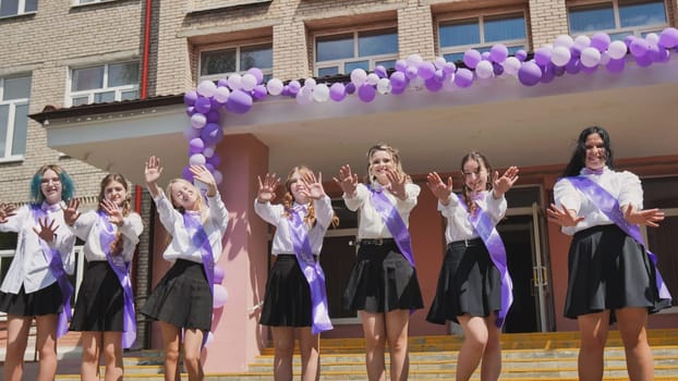 Group of cheerful female graduates waving goodbye on their last school day, celebrating the end of academic year with purple ribbons and balloons