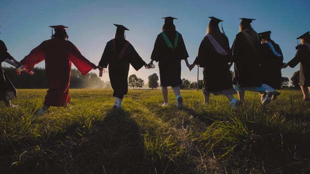 Group of students wearing graduation gowns walking together in a field at sunset, holding hands and celebrating their achievement