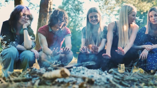 Five young students are crouching around a campfire, warming their hands and enjoying their time together in nature