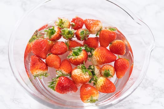Ripe strawberries are submerged in water within a large glass mixing bowl, a step in washing the fruit to ensure cleanliness and longevity before storage or consumption.