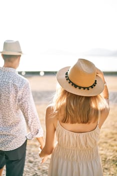 Woman follows man along the beach, holding his hand and holding her hat. Back view. High quality photo