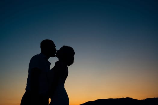 Silhouettes of kissing bride and groom against the backdrop of a sunset over the mountains. High quality photo