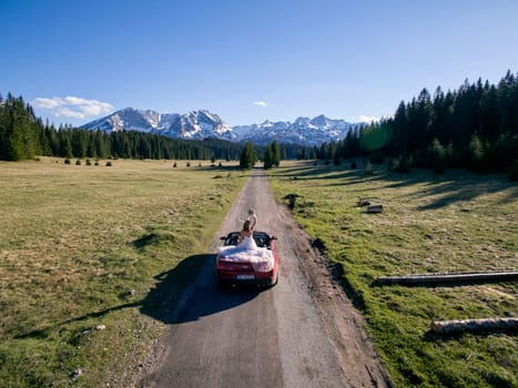 Bride with a bouquet stands in a cabriolet driving along the road in a mountain valley. Back view. Drone. High quality photo