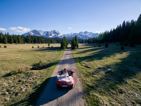 Bride stands in a cabriolet driving along the road in a mountain valley. Back view. Drone. High quality photo