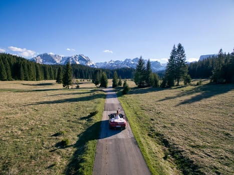 Bride waves her bouquet while sitting in a cabriolet driving along a high mountain road. Back view. Drone. High quality photo