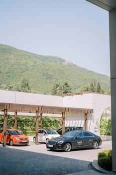 Multi-colored cars stand in the hotel parking lot under a wooden canopy at the foot of the green mountains. High quality photo