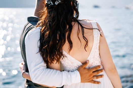 Bride and groom stand hugging and looking at the sea. Back view. Cropped. High quality photo