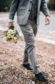 Groom with a bouquet of flowers in his hand walks along the pebble beach. Cropped. Faceless. High quality photo