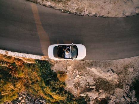 Bride and groom are driving along the highway on a slope in a white convertible. Drone. High quality photo