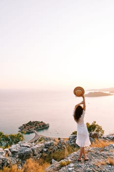 Woman with a hat raised above her head stands on a mountain and looks at the sea. Sveti Stefan, Montenegro. Back view. High quality photo