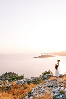 Girl with a hat in her hands above her head stands on a mountain above the island of Sveti Stefan. Montenegro. Back view. High quality photo