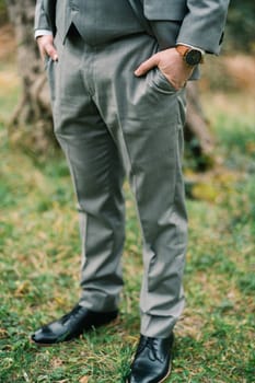 Groom with his hands in his pants pockets stands on the green grass. Cropped. Faceless. High quality photo