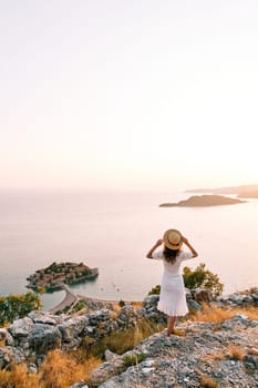 Girl straightens her hat standing on the mountain and looks at the island of Sveti Stefan. Montenegro. Back view. High quality photo