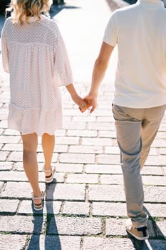 Man and woman walk holding hands along a sunny paved road. Back view. Cropped. High quality photo