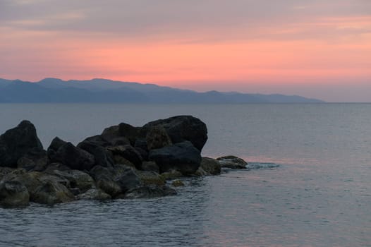 Soft pastel colors fill the sky as the sun sets over calm waters, with rugged rocks jutting into the serene landscape, creating a peaceful atmosphere for reflection and relaxation.