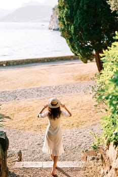 Young woman in a sundress goes to the beach, holding her hat on her head. Back view. High quality photo