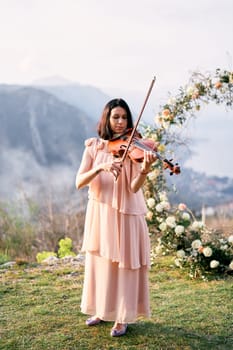 Girl musician plays the violin near the wedding arch on the mountain. High quality photo