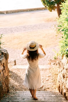 Girl goes down the stone steps to the beach, touching the hat on the head. Back view. High quality photo