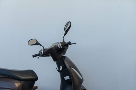 A sleek, brown motorcycle stands idle beside a simple white wall, symbolizing urban mobility and freedom. The shiny mirrors reflect a tranquil moment in a bustling city environment.