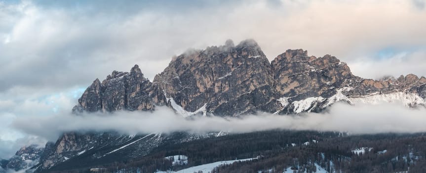 Aerial view of rugged Dolomite peaks near Cortina d'Ampezzo, Italy, with snow covered slopes, low clouds, and visible ski trails and structures.