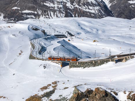 An orange train travels on a curved railway bridge of the Gornergrat Railway in snowy Zermatt, Switzerland, with rugged peaks in the background.