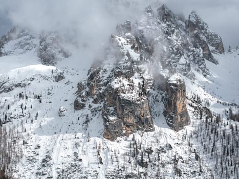 A dramatic snow covered mountain in the Dolomites near Cortina, Italy, with rugged peaks, sparse trees, and mist partially shrouding the cliffs.