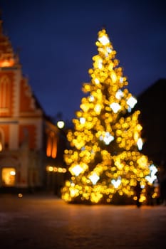A majestic Christmas tree adorned with sparkling lights stands proudly in a vibrant square as the sun sets, casting a warm golden glow across the bustling holiday scene.