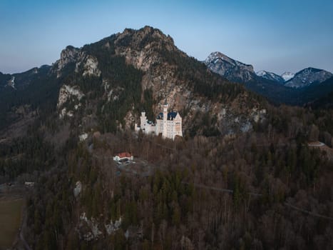 Aerial view of Neuschwanstein Castle in Bavaria, Germany, surrounded by dense woodland and rugged mountains with snow capped peaks in the distance.