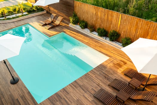 Modern turquoise swimming pool with wooden deck, lounge chairs, and umbrellas in a stylish backyard