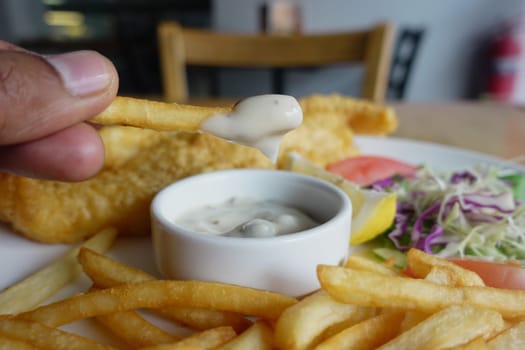 A hand holds a fry dipped in sauce, surrounded by a delicious meal.