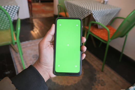 A hand displays a smartphone featuring a blank green screen inside a cafe.