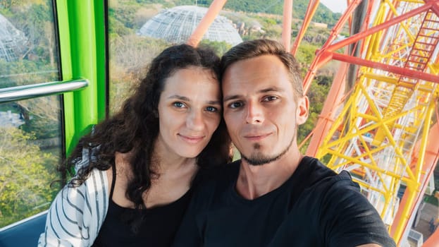 Selfie of happy couple tourists travelers in love in cabin of a ferris wheel in amusement park on summer vacation