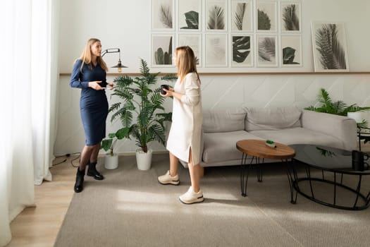 In a contemporary living room, two women discuss animatedly. One wears a fitted dress and the other opts for a casual outfit, both surrounded by greenery and elegant decor.