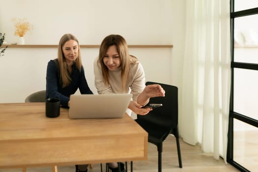 Two women engage in a collaborative discussion at a wooden table in a well-lit workspace. They review content on a laptop while exchanging ideas.