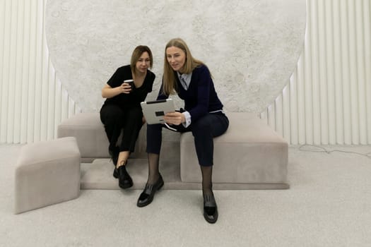 Two women are engaged in a work-related discussion while using a digital device. They are seated on a stylish sofa in a contemporary, minimalistic environment.