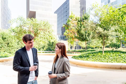 smiling man and a woman standing in a park near of office buildings talking while taking coffee, they are smiling and enjoying with the company, concept of business, copyspace for text