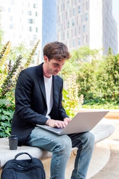 smiling young man using a laptop sitting on a bench of a park near of office buildings, concept of business and technology for remote work