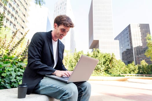 smiling young man using a laptop sitting on a bench of a park near of office buildings, concept of business and technology for remote work